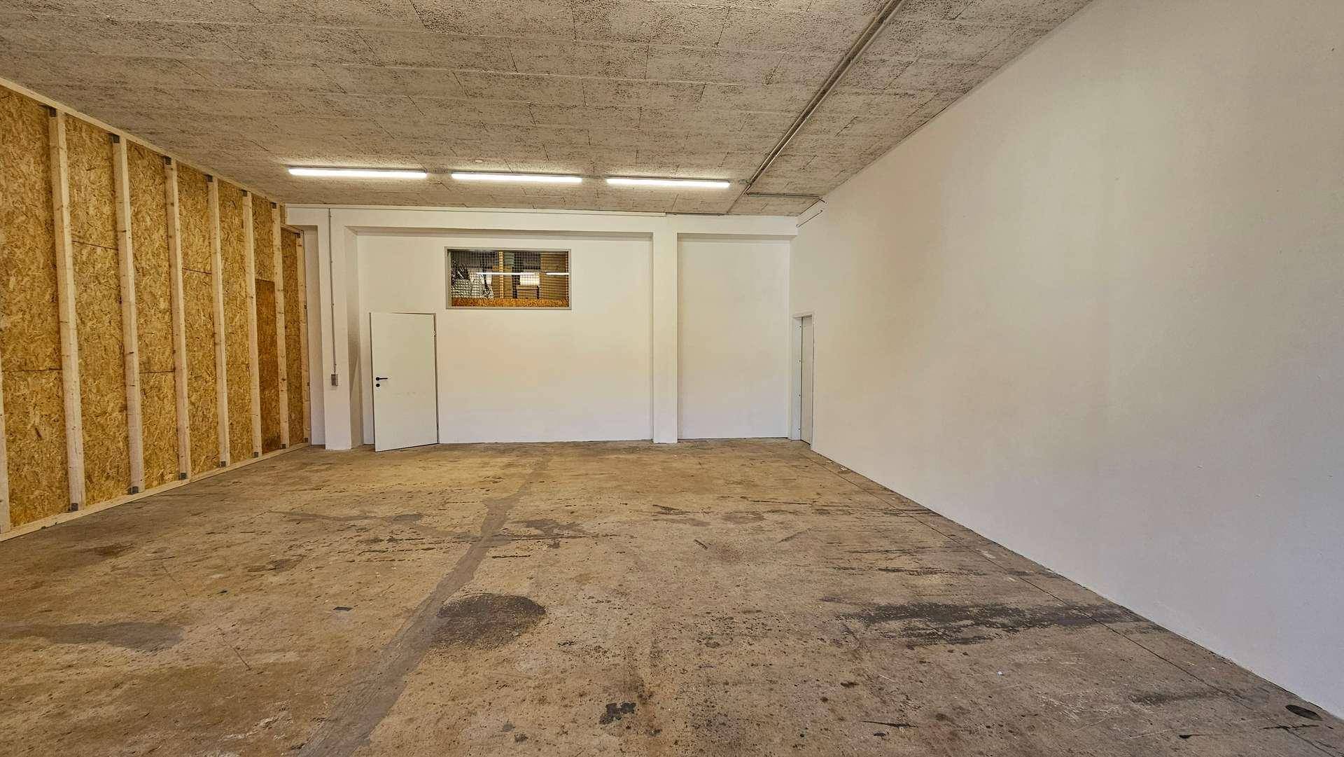 Leerer Gewerberaum mit Betonboden, Holzverkleidung, einer Tür und einem kleinen Fenster, bereit zur Nutzung.