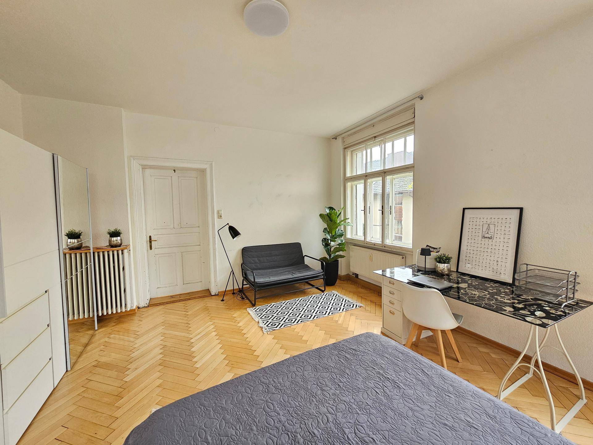 Schlafzimmer mit Fischgrätparkett, weißer Tür und einem kleinen Sofa neben dem Fenster.