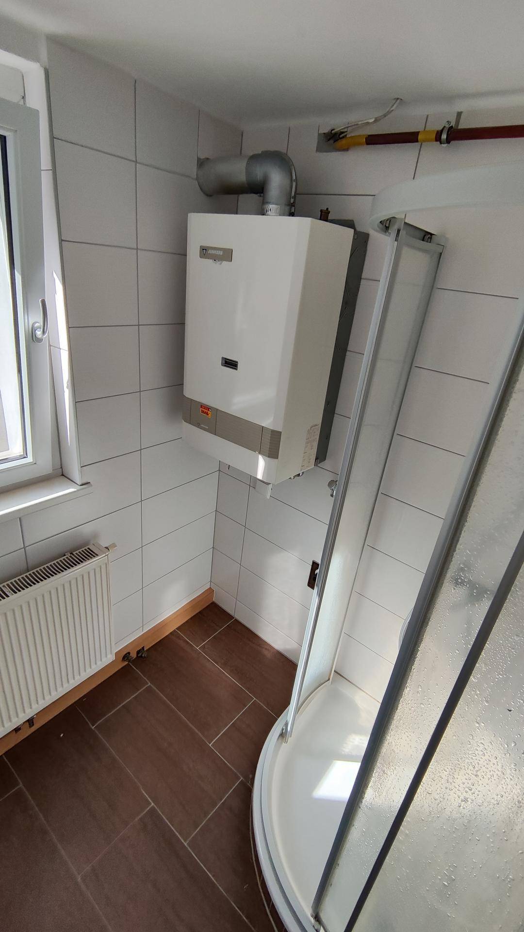 Badezimmer mit Duschkabine, Fenster und einem Warmwasserbereiter an der Wand.