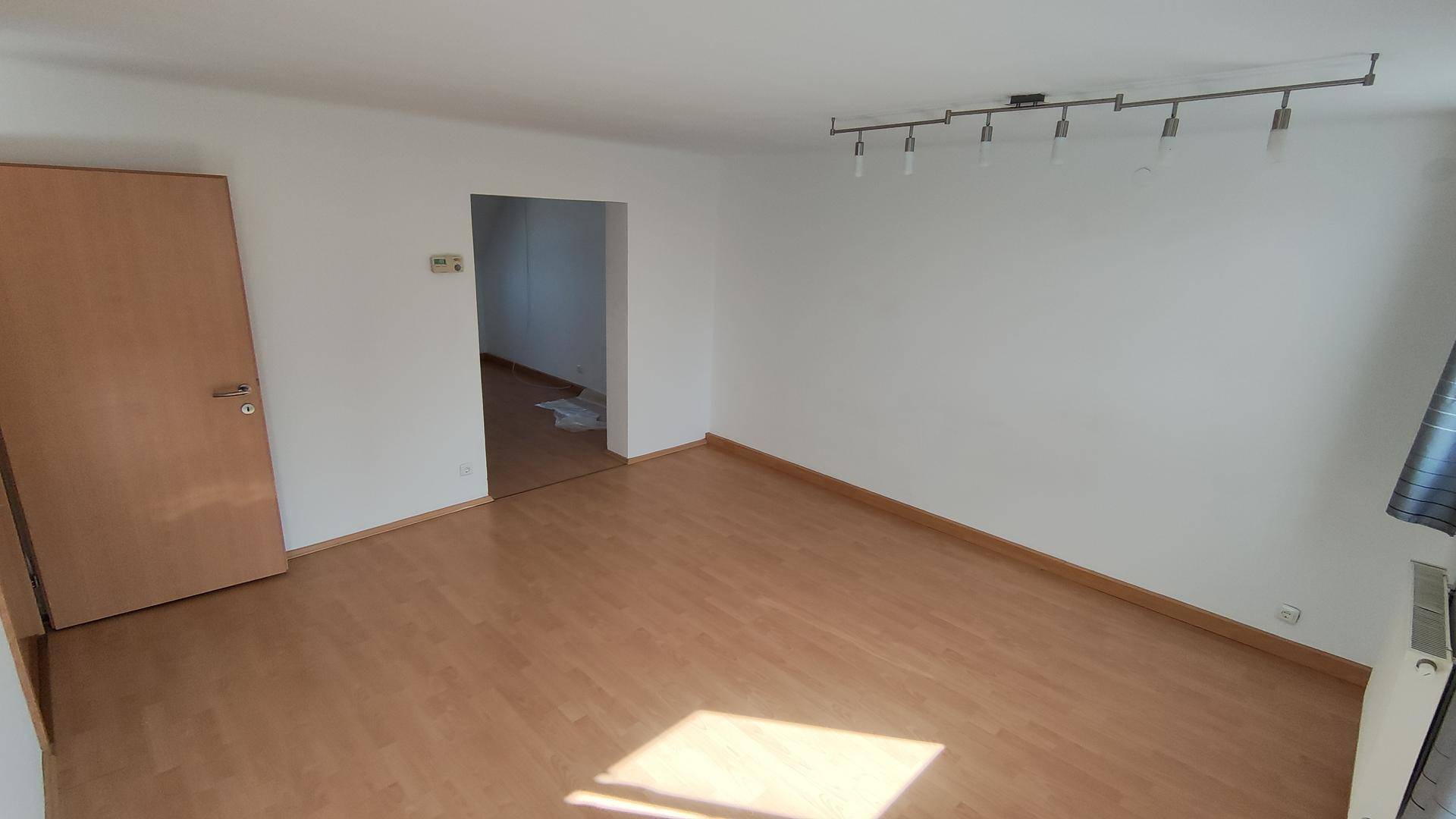 Leeres Wohnzimmer mit hellem Laminatboden, weißen Wänden und Zugang zu einem weiteren Raum.