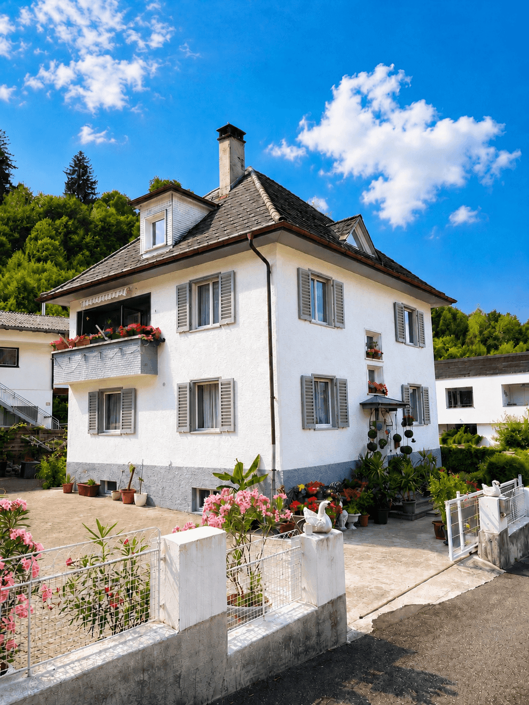 Helles Einfamilienhaus mit traditioneller Architektur, Mansarddach und üppiger Bepflanzung im Vorgarten.