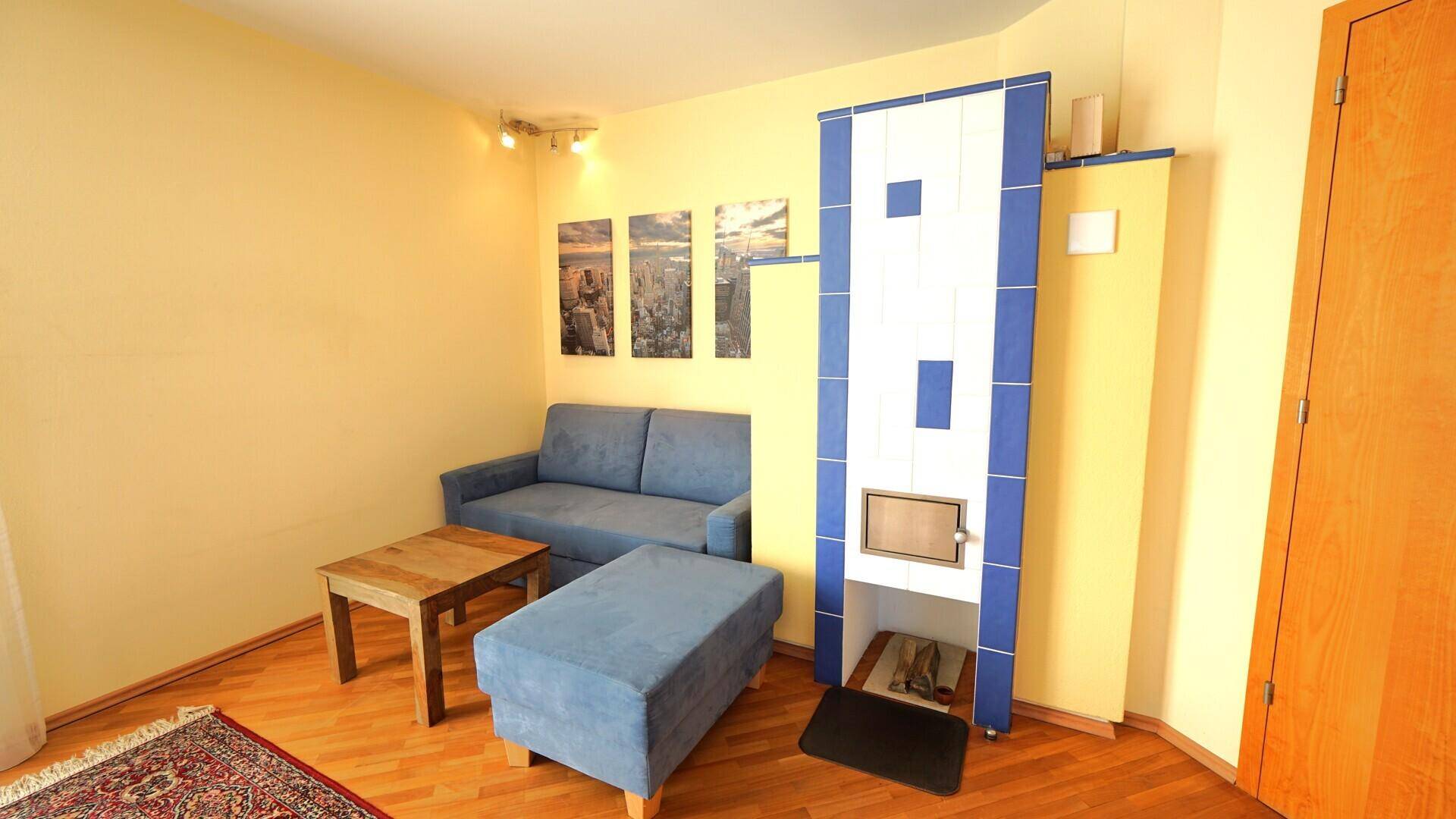 Gemütliches Wohnzimmer mit blauem Sofa, Holzboden und einem traditionellen Kachelofen.