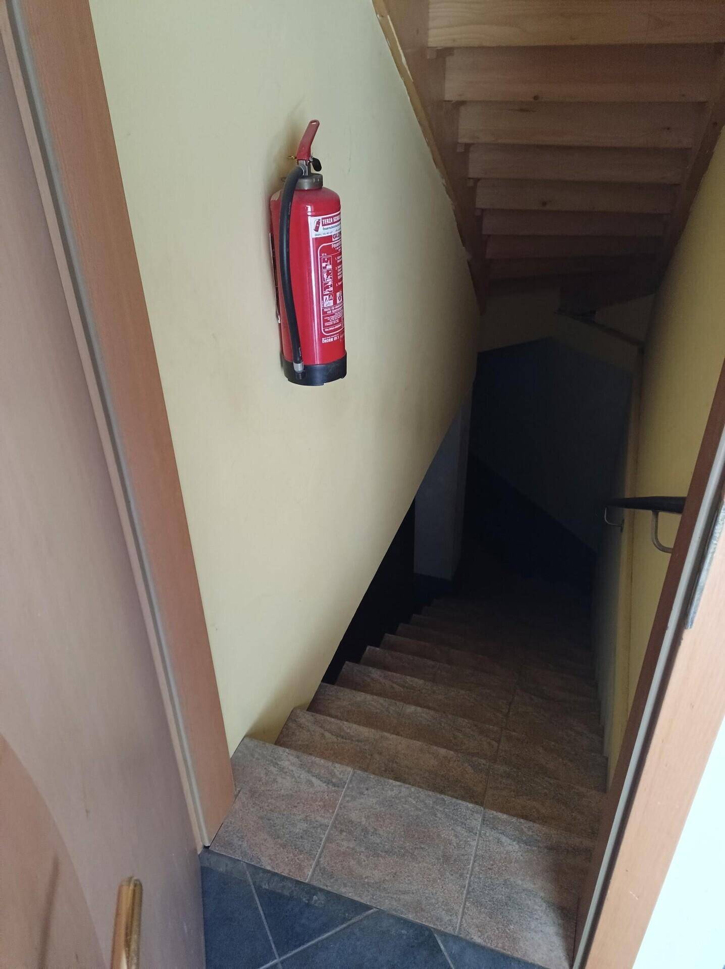 Treppenabgang in den Keller mit Fliesenboden und einem Feuerlöscher an der Wand.