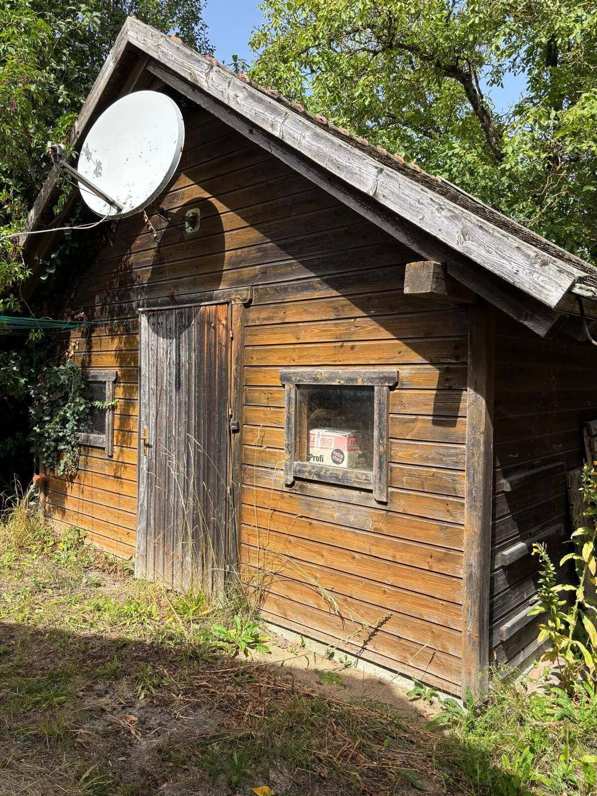 Holzschuppen mit Satellitenschüssel und einfacher Holztür in ländlicher Umgebung.