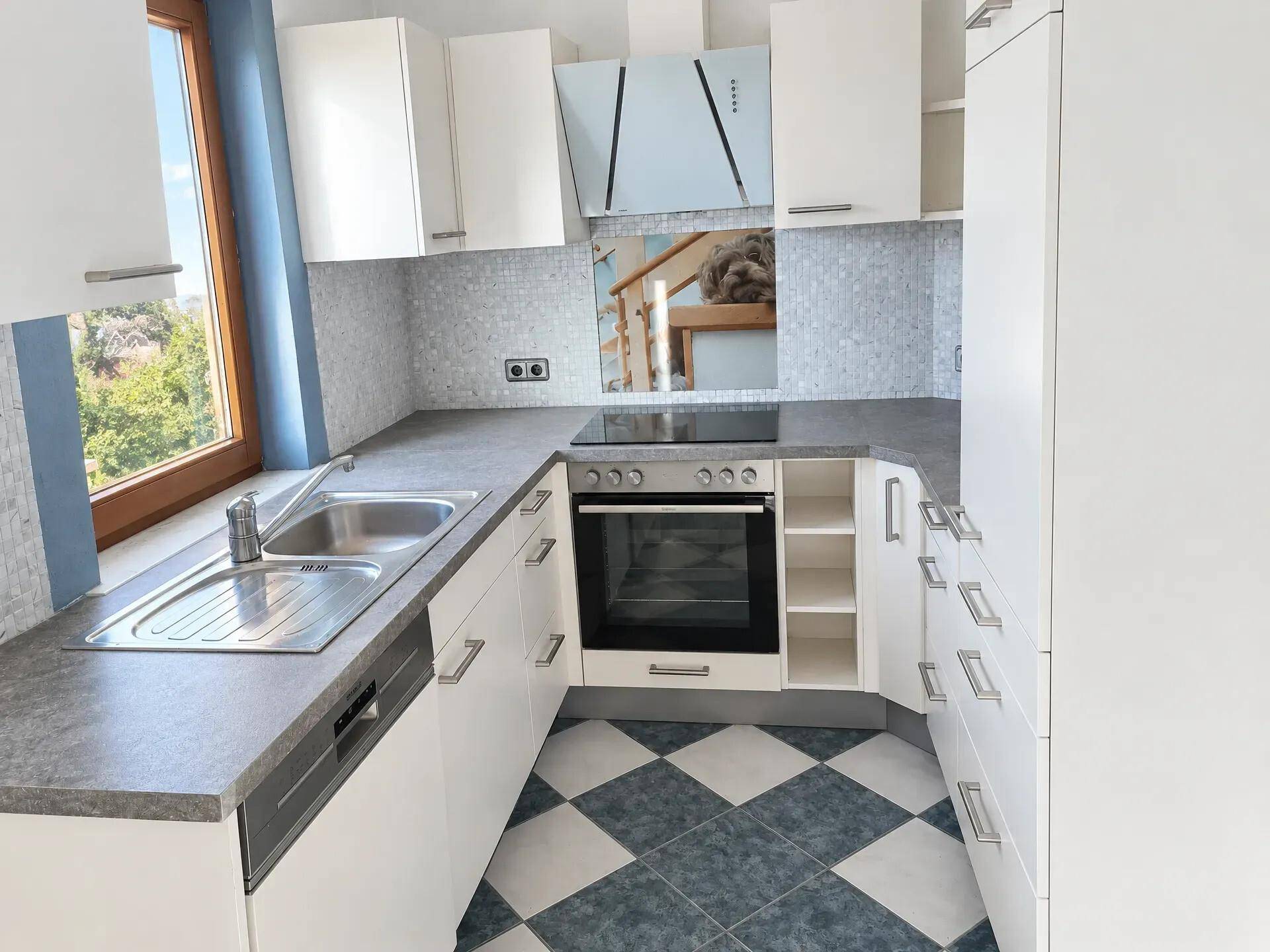 Moderne Küche mit weißen Schränken, grauer Arbeitsplatte, Einbaugeräten und karierten Bodenfliesen.