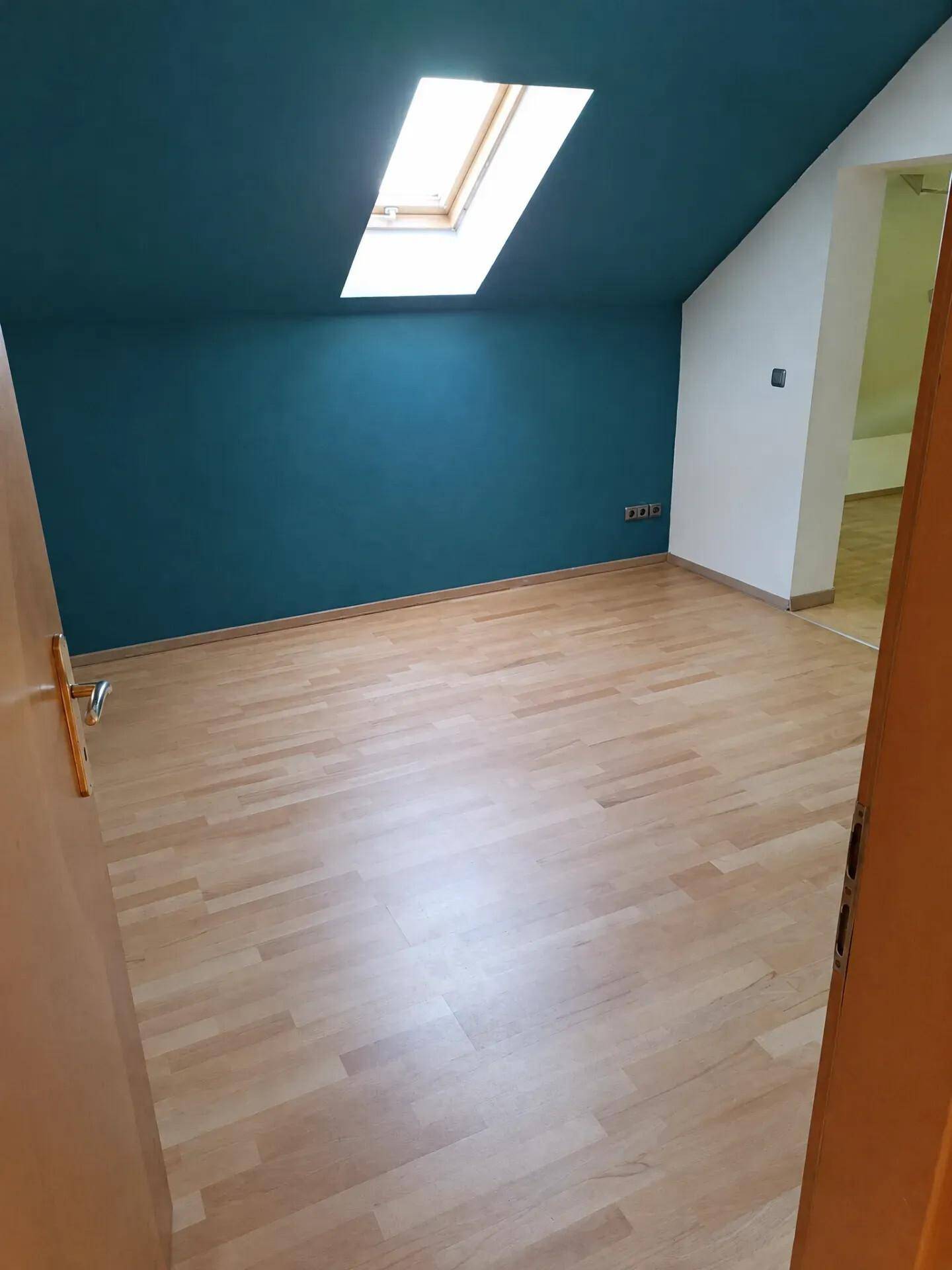 Helles Zimmer mit Dachschräge, Dachfenster, blauer Akzentwand und Holzboden.