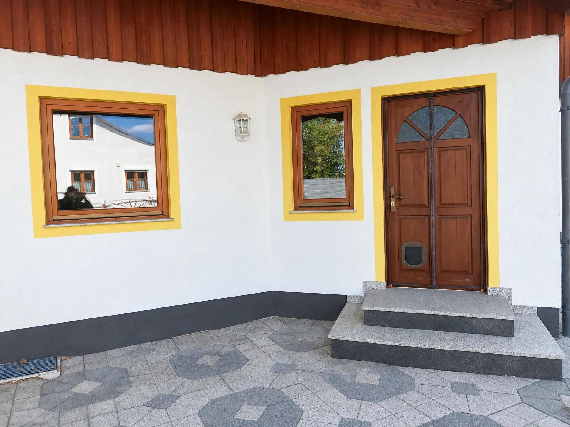 Hauseingang mit Holztür, gelben Fensterrahmen und einem gepflasterten Vorplatz in gutem Zustand.