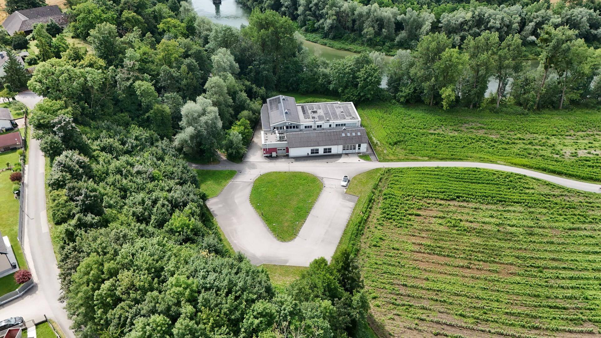 Weitläufige Luftaufnahme des Industriegebäudes inmitten von Feldern, Bäumen und einem Fluss im Hintergrund.