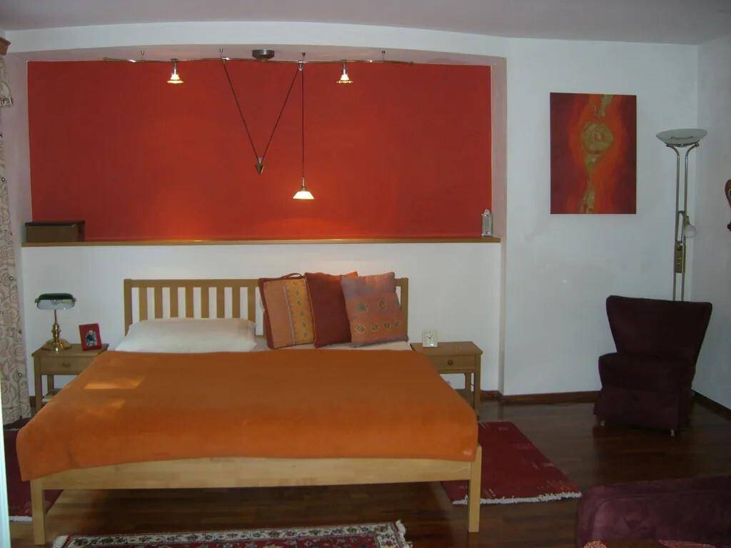 Schlafzimmer mit orangefarbener Bettwäsche, roter Akzentwand und Holzboden.