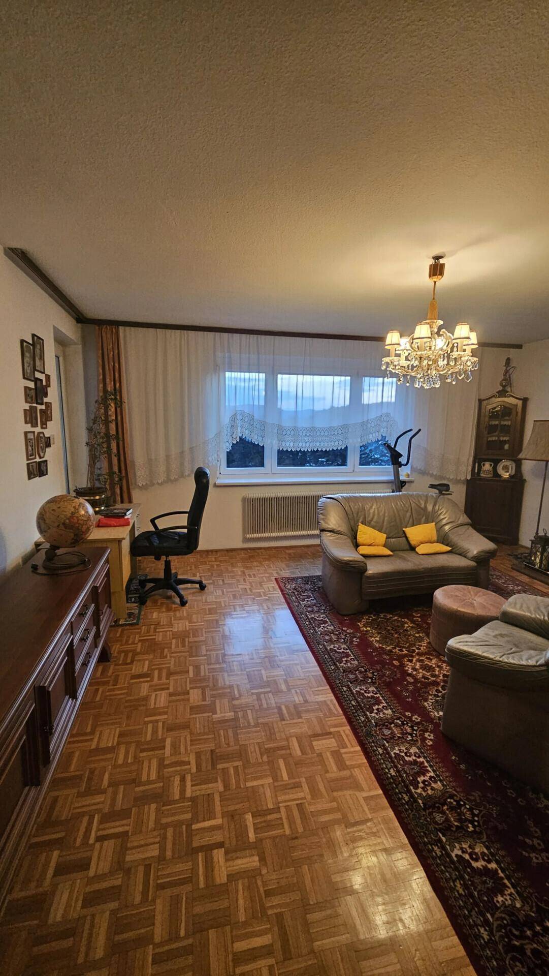 Wohnzimmer mit Parkettboden, Schreibtischbereich und großem Fenster am Abend.