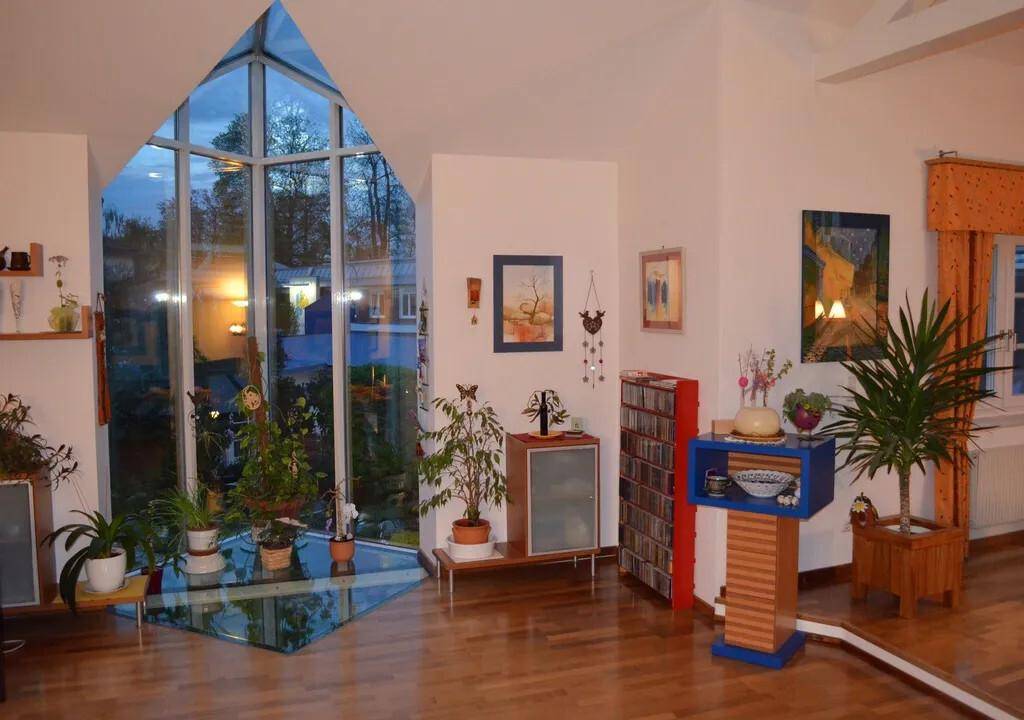 Geräumiges Wohnzimmer mit großen Fenstern, Holzboden und vielen Pflanzen.