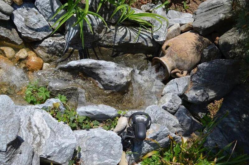 Kleiner Wasserfall mit Steinen und einer antiken Amphore als Dekoration im Garten.