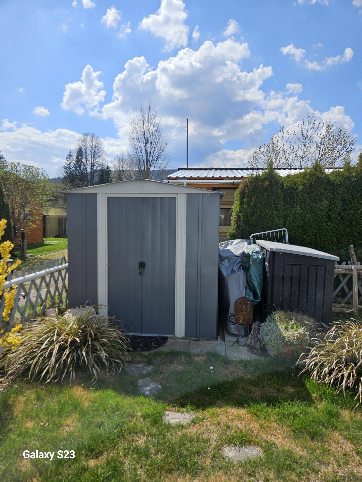 Grauer Gartenschuppen im Grünen, umgeben von Pflanzen und Sträuchern unter blauem Himmel.