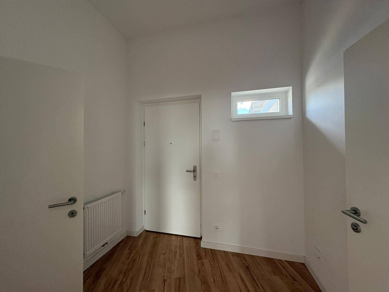 Heller Flur mit Eingangstür, kleinem Fenster und Heizkörper, ausgestattet mit Laminatboden.