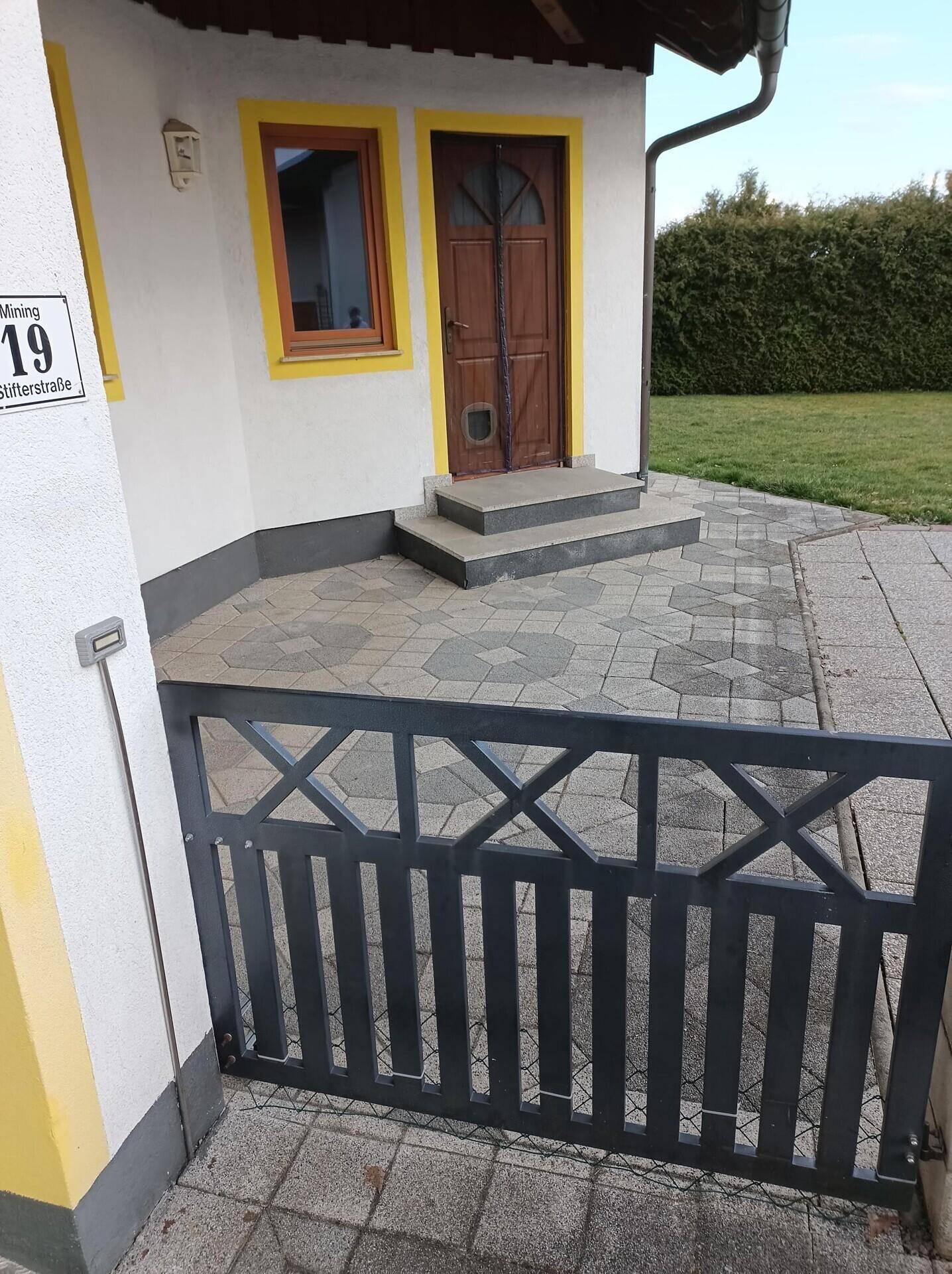 Eingangsbereich des Hauses mit gepflastertem Weg, Zaun und Hausnummer.