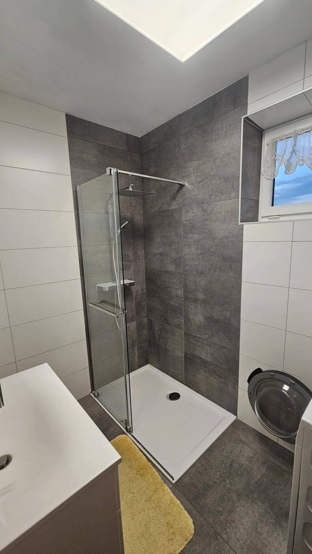 Helles Badezimmer mit moderner ebenerdiger Dusche, Fenster und grauen Fliesen.