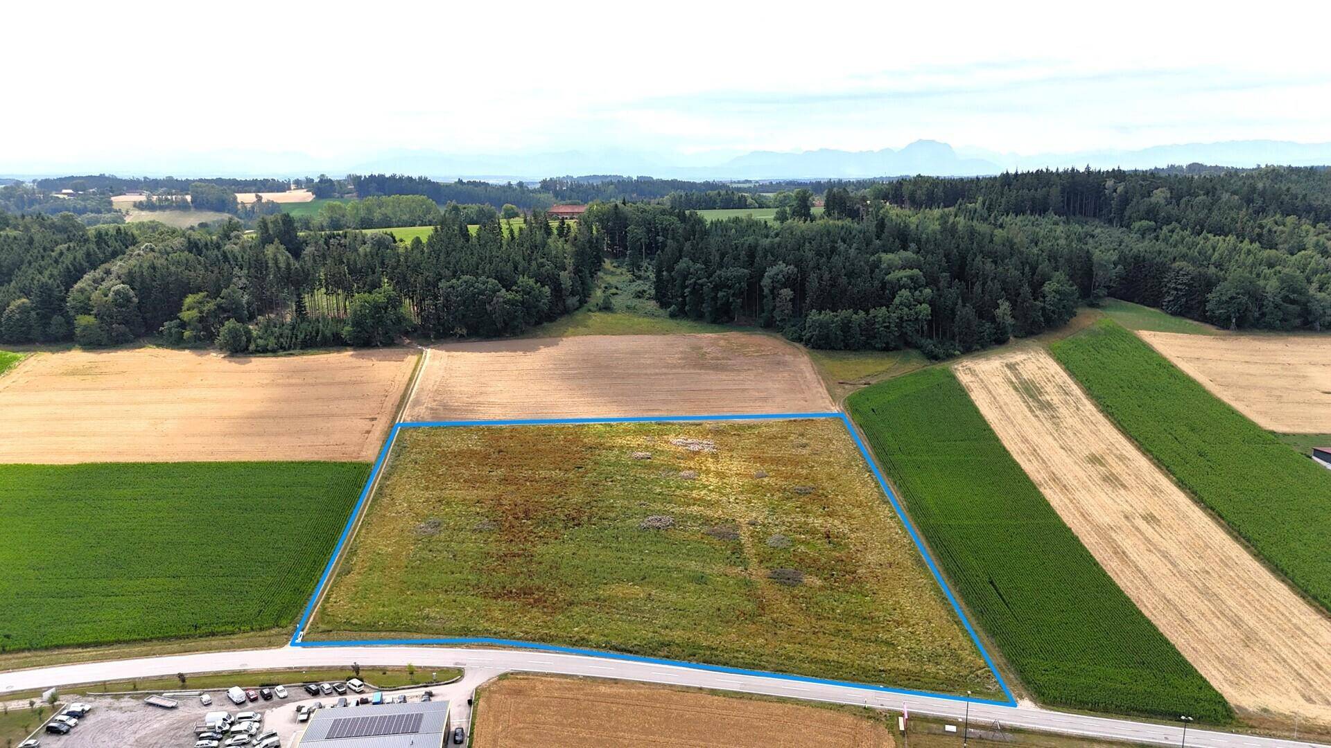 Luftaufnahme eines großen, blau umrandeten Grundstücks, umgeben von Feldern und Wäldern mit Bergen im Hintergrund.