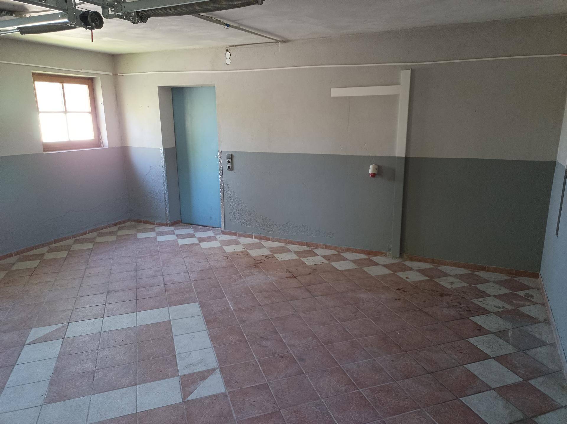 Geräumige Garage mit Fliesenboden, Fenster und Zugang zu einem weiteren Raum.