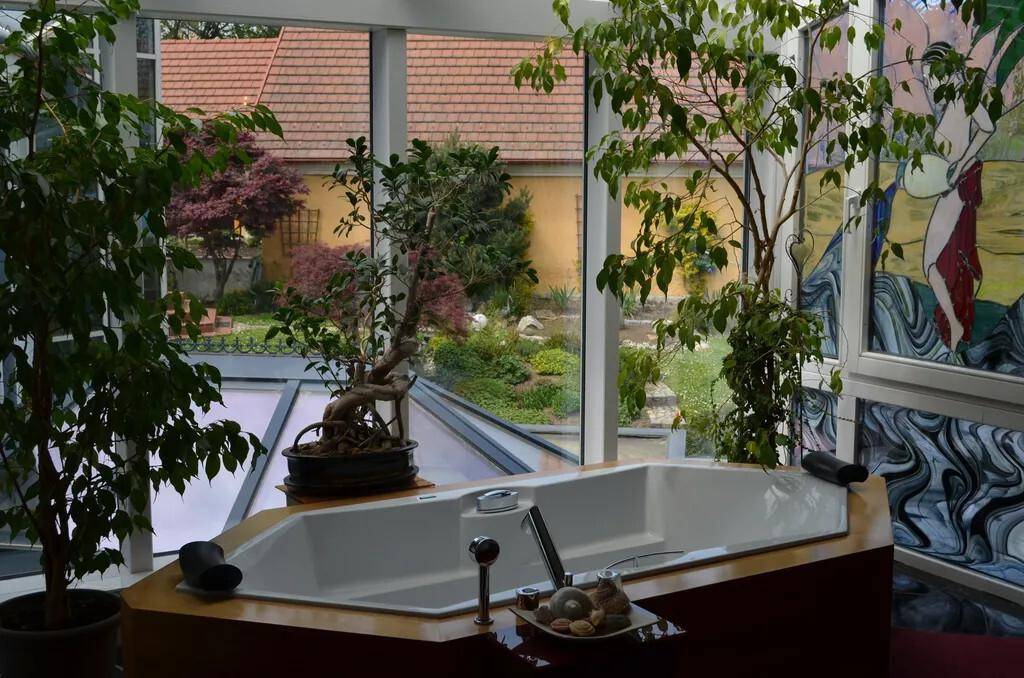 Badezimmer mit Badewanne in einem Wintergarten, umgeben von Pflanzen und Buntglasfenstern.