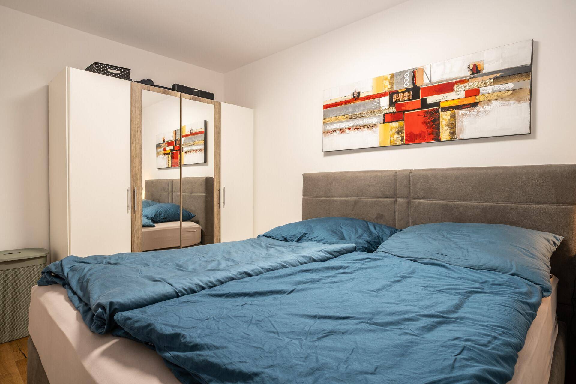 Helles Schlafzimmer mit Doppelbett, Spiegelschrank und modernem Kunstwerk an der Wand.
