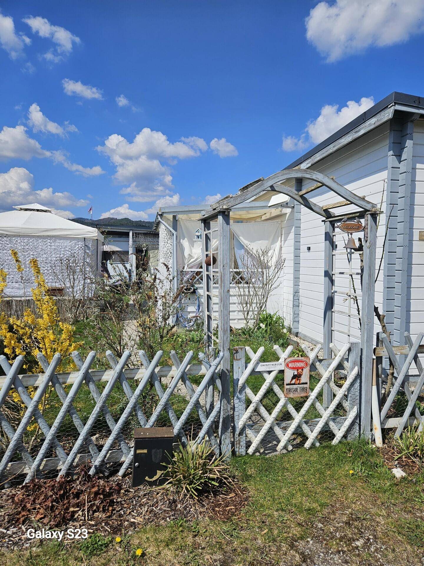 Vorderansicht eines kleinen weißen Hauses mit Gartenzaun und blühenden Sträuchern.
