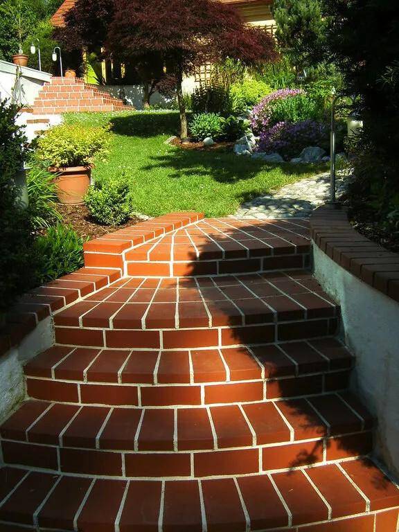 Geschwungene Ziegelsteintreppe führt durch den sonnigen Garten mit Blumenbeeten.
