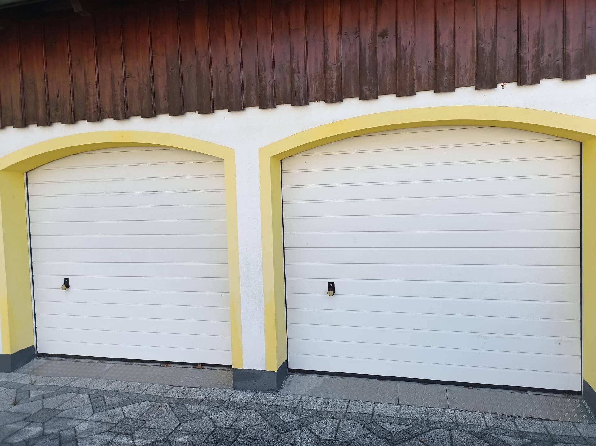 Zwei weiße Garagentore mit gelben Bögen unter einer Holzfassade und gepflastertem Vorplatz.