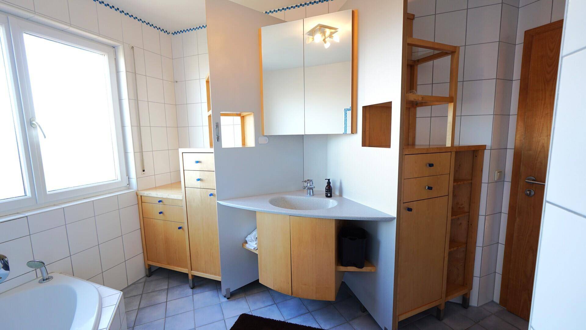 Badezimmer mit Eckbadewanne, Einzelwaschbecken und Spiegelschrank, hell gefliest.
