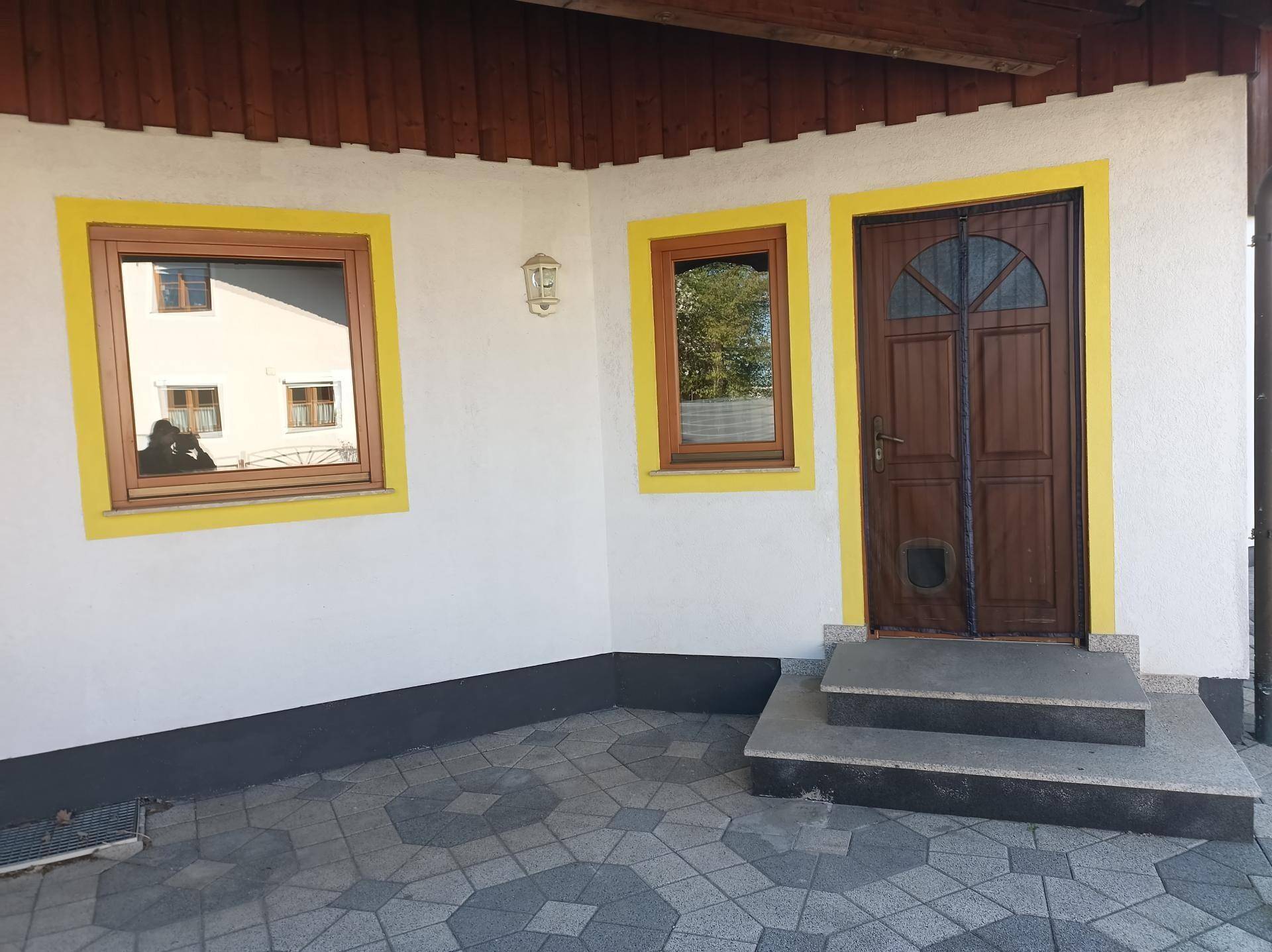 Eingangsbereich eines Hauses mit gelb umrahmten Fenstern und einer Holztür, davor ein gepflasterter Bereich.