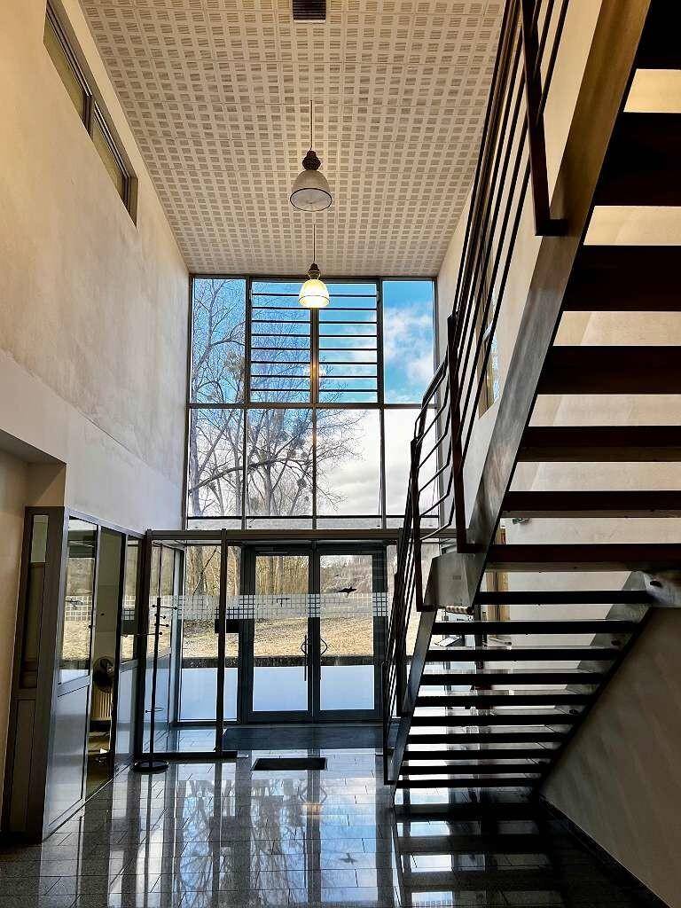 Heller Eingangsbereich mit hoher Decke, großen Glasfronten und einer modernen Treppe.