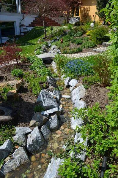Naturnaher Bachlauf mit Steinen und üppiger Bepflanzung in einem terrassierten Garten.