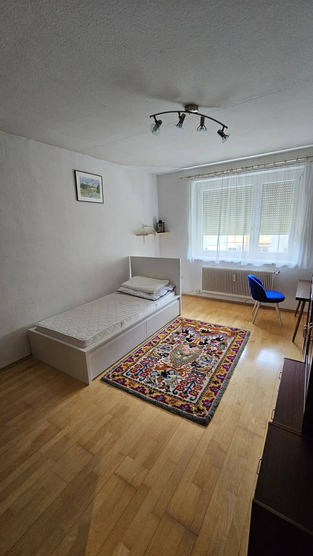 Helles Schlafzimmer mit Einzelbett, Holzboden und einem bunten Teppich.