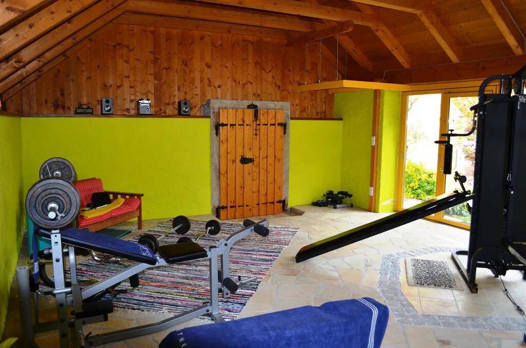 Fitnessraum mit Trainingsgeräten, Holzbalkendecke und farbigen Wänden.