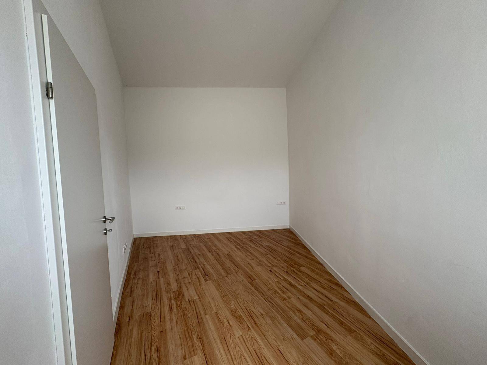 Schlichtes Zimmer mit weißen Wänden und hellem Laminatboden, bietet vielseitige Nutzungsmöglichkeiten.