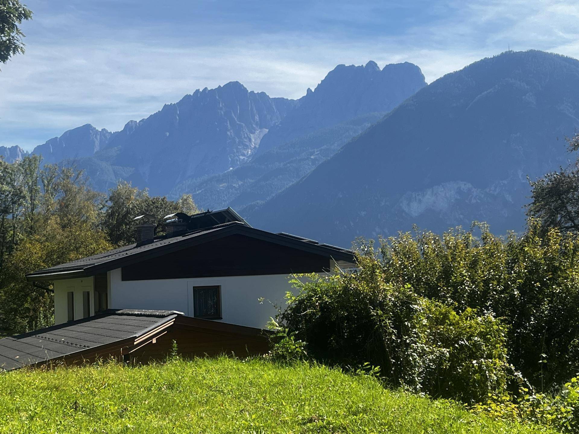 Modernes Haus mit dunklem Dach und weißer Fassade, eingebettet in grüne Landschaft mit Bergblick.