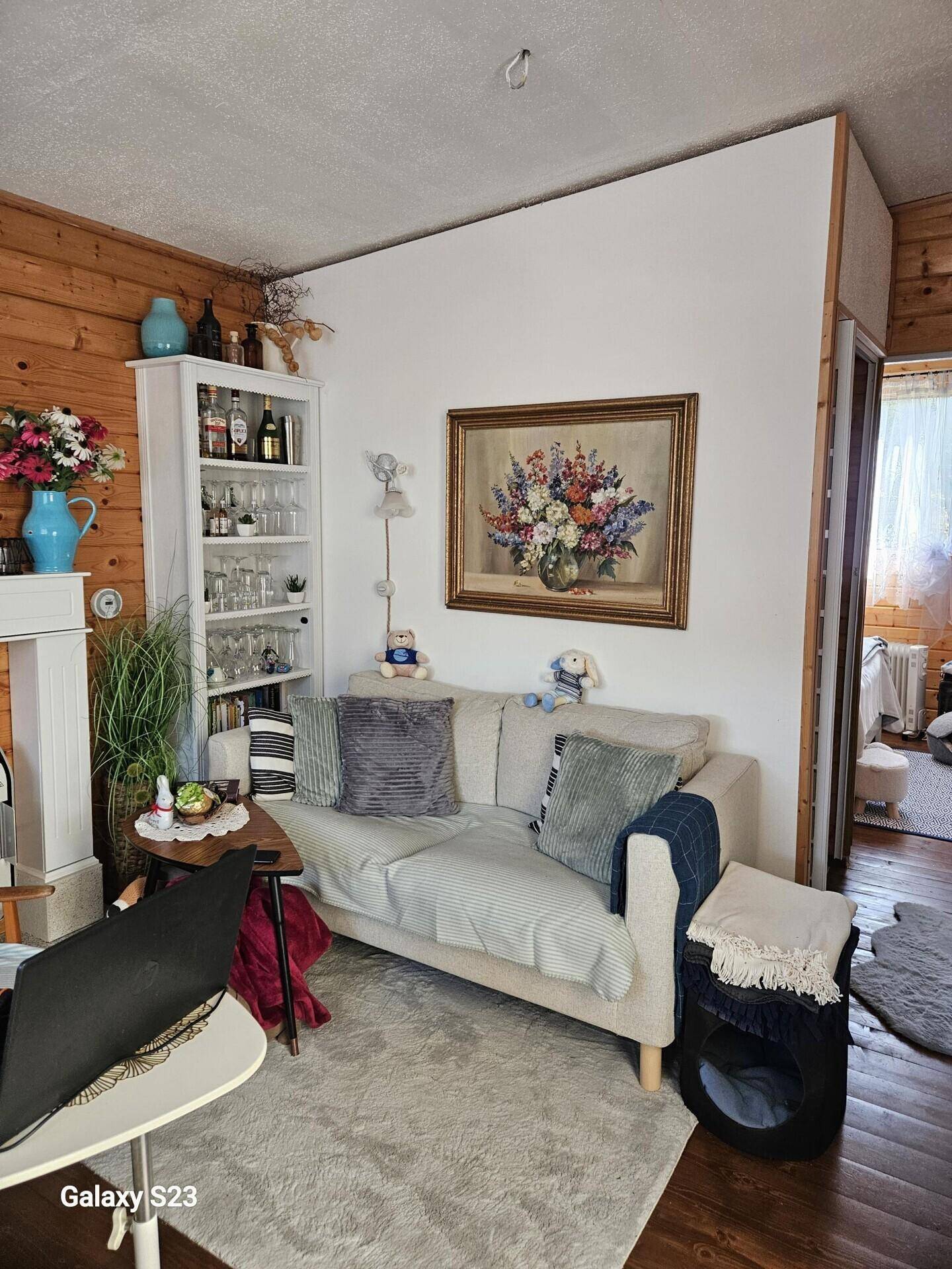 Wohnzimmer mit Holzwänden, Sofa, Bücherregal und dekorativen Blumenarrangements.
