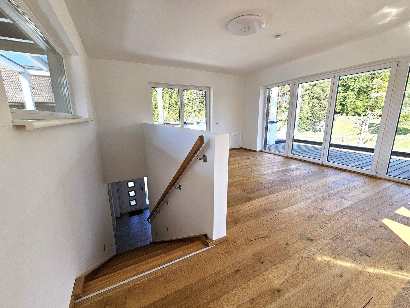 Heller Flur im Obergeschoss mit Holzparkett, Treppe und großen Fenstern mit Waldblick.