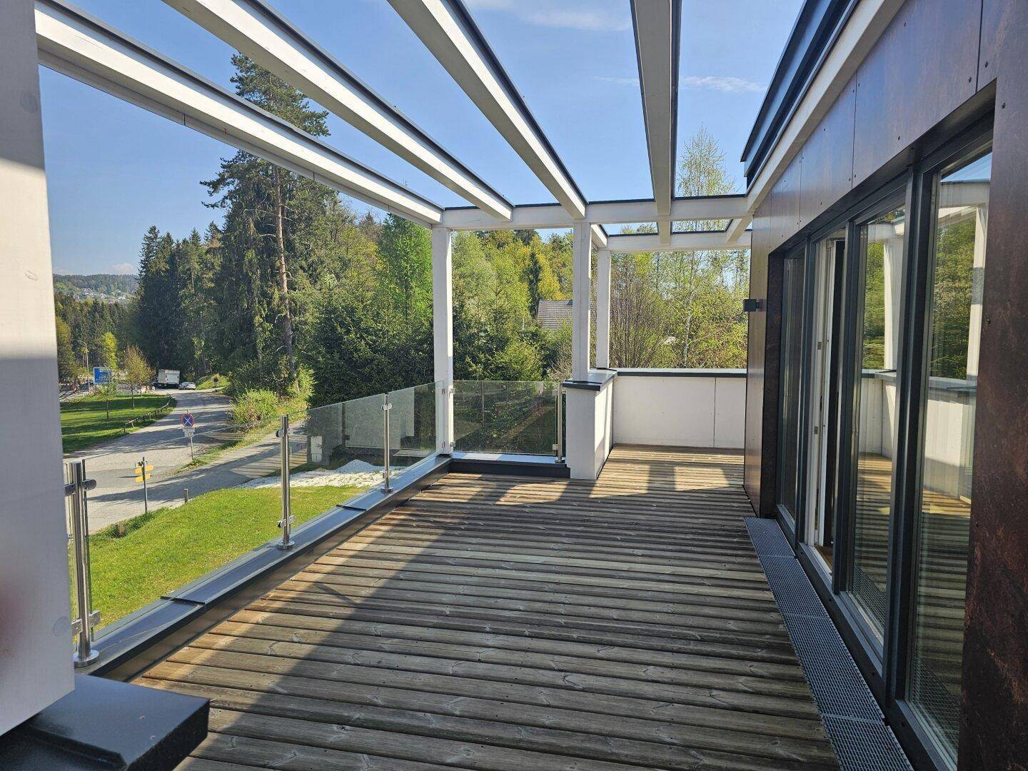 Große Holzterrasse mit Pergola-Struktur und Glasgeländer, umgeben von viel Grün.