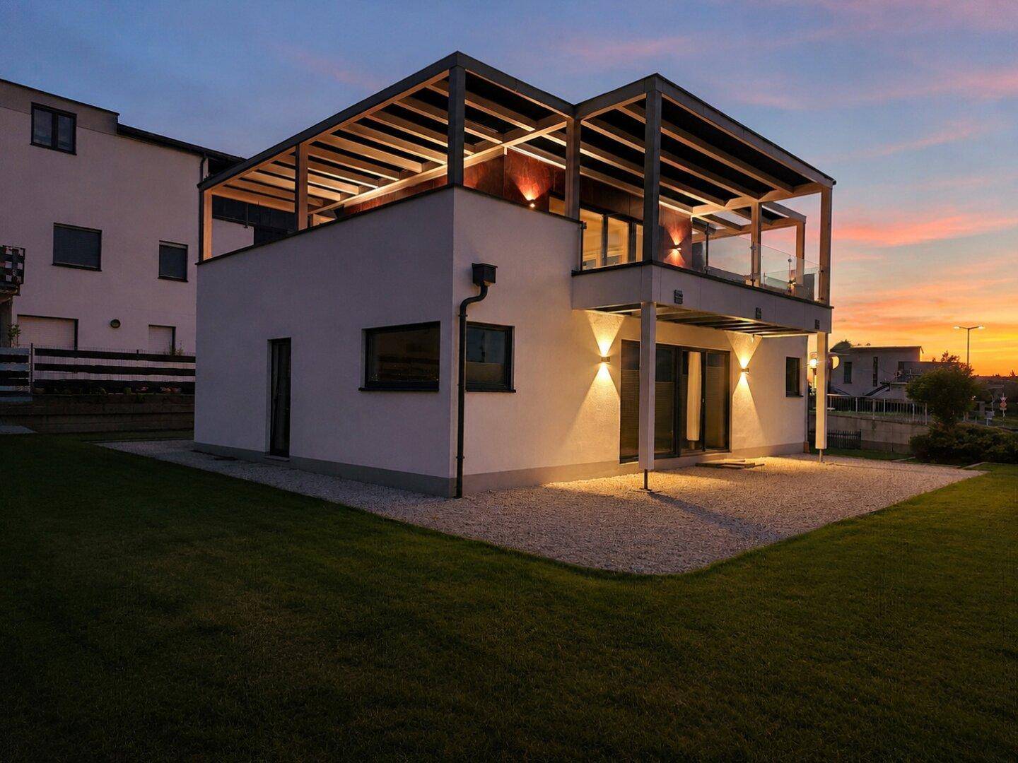 Seitenansicht des modernen Hauses mit beleuchteter Terrasse und gepflegtem Rasen bei Sonnenuntergang.