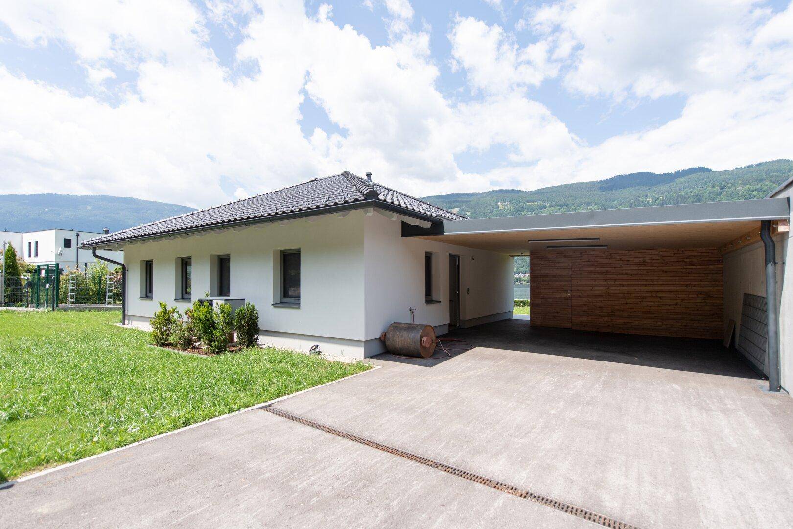 Modernes Haus mit Carport und Zufahrt, umgeben von Rasen und Bergen.