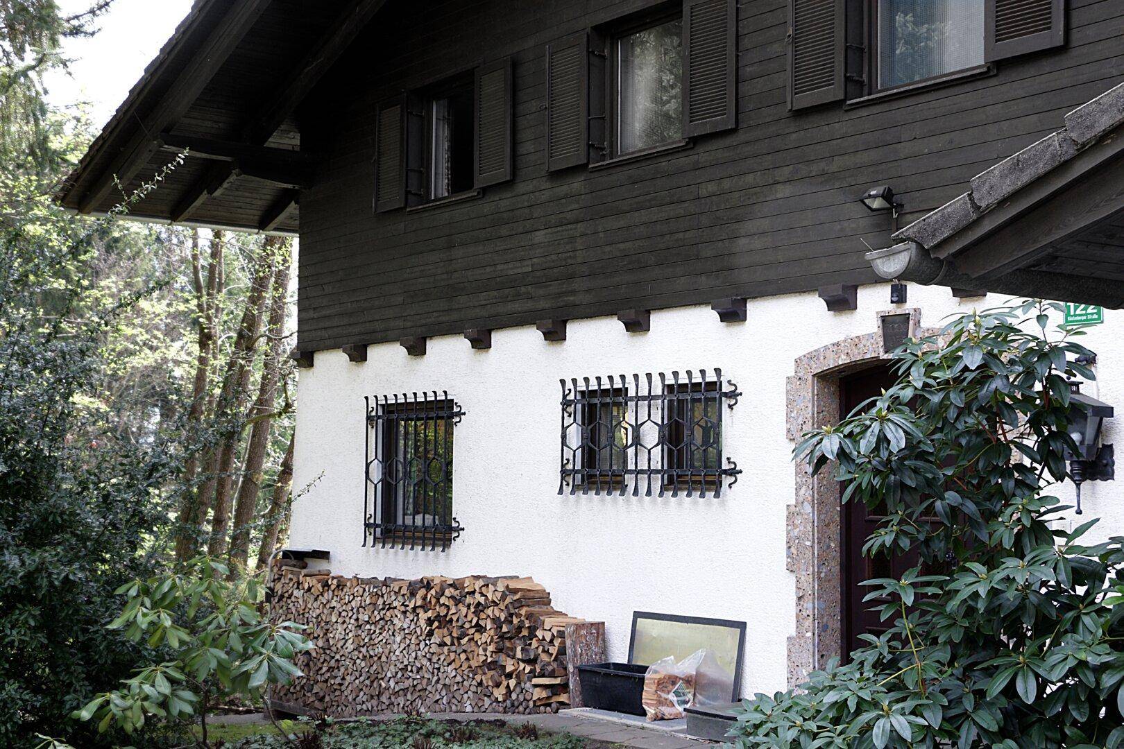 Detailansicht der Hausfassade mit weißem Putz, dunkler Holzverkleidung, Fensterläden und einem Holzstapel.