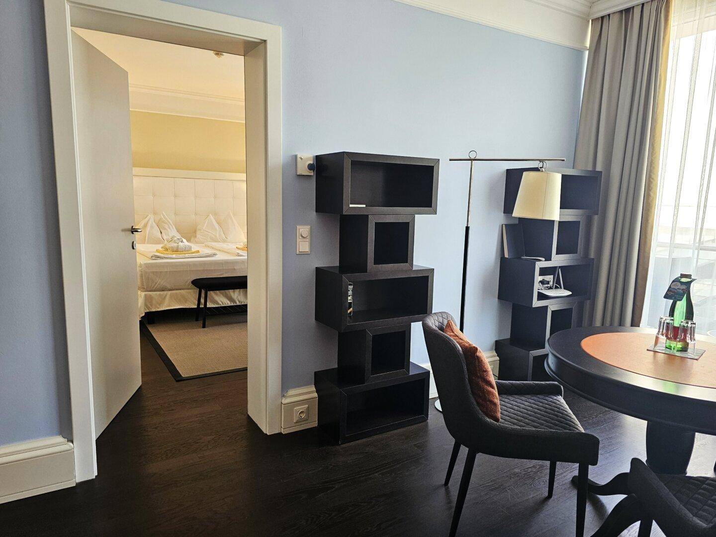 Sitzbereich mit dunklem Holzregal und Blick in ein helles Schlafzimmer mit Doppelbett.