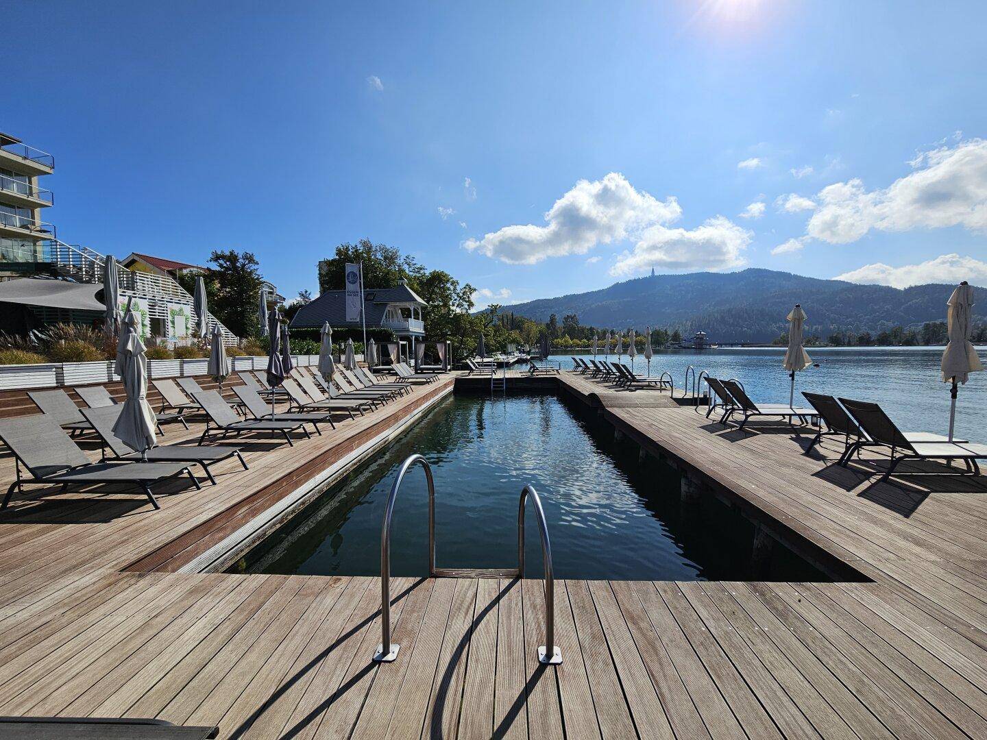 Außenpool auf einer Holzterrasse direkt am See mit Liegestühlen und Bergblick.