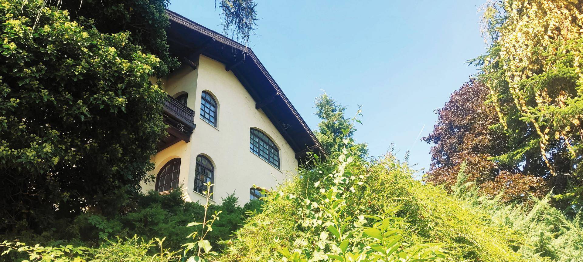 Hausfassade mit Rundbogenfenstern, umgeben von dichtem Baumbestand und Vegetation.