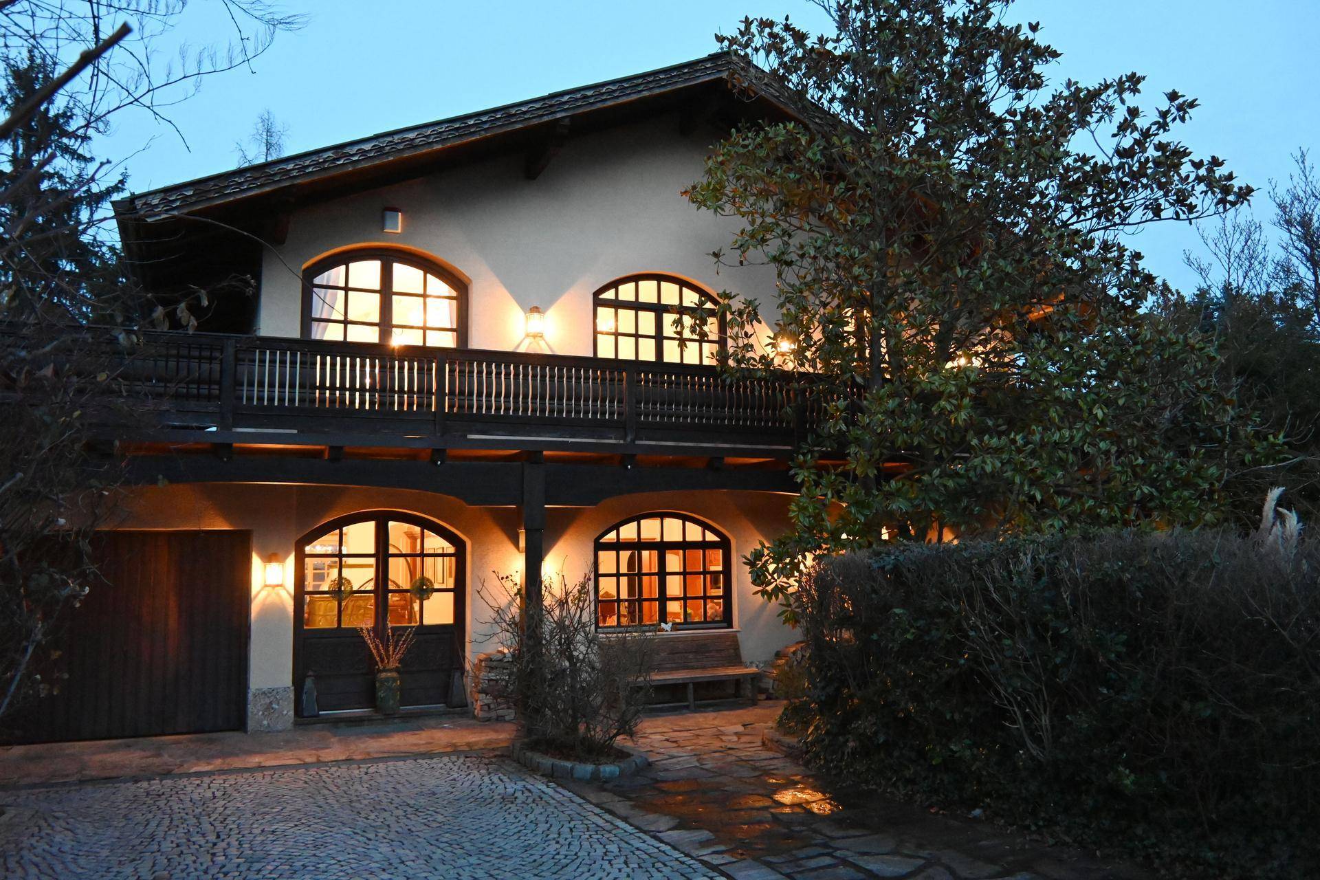 Beleuchtete Hausfassade in der Dämmerung mit Balkon und gepflasterter Auffahrt.