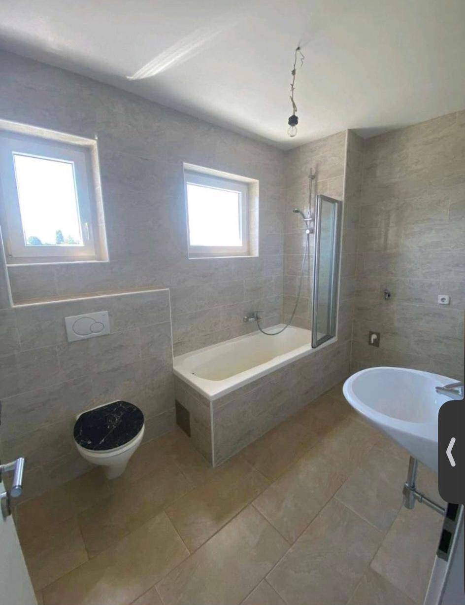 Modernes Badezimmer mit hellgrauen Fliesen, Badewanne mit Duschwand, WC und Waschbecken.