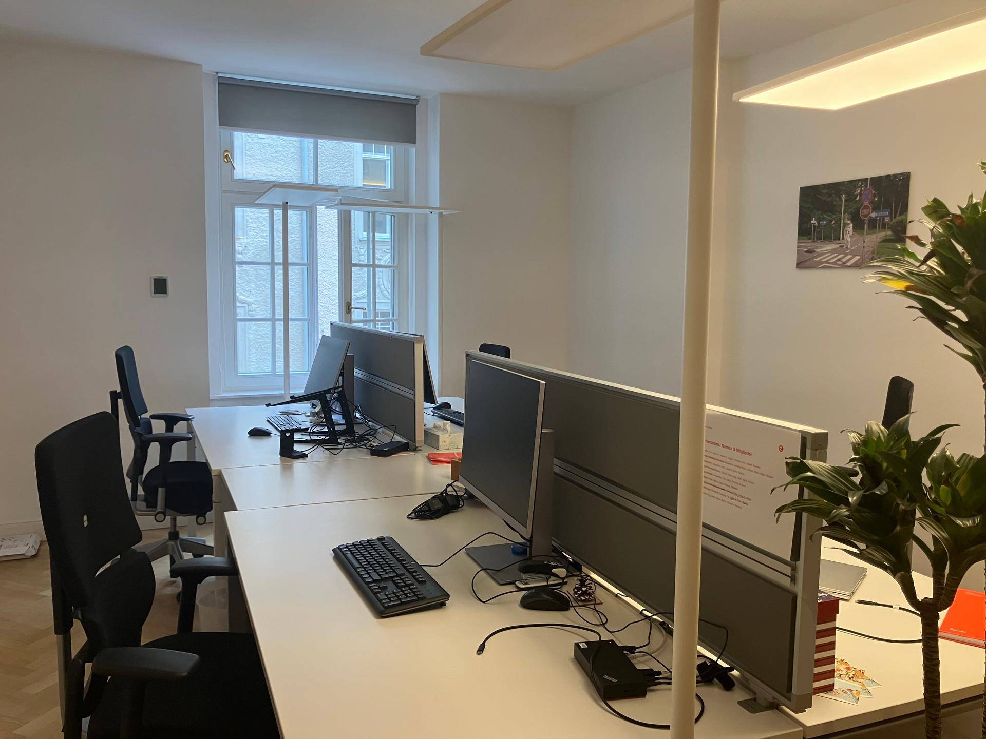 Heller Büroraum mit mehreren Arbeitsplätzen, Computern und einem Fenster mit Blick nach draußen.