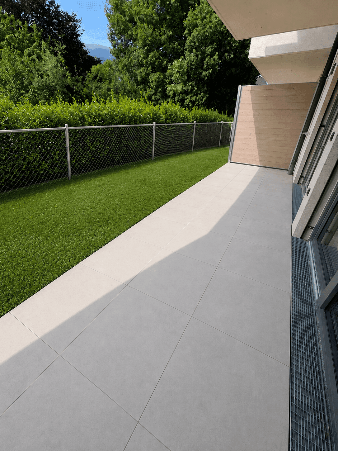 Moderne Terrasse mit hellen Fliesen und angrenzendem Rasenbereich, umgeben von einer grünen Hecke.