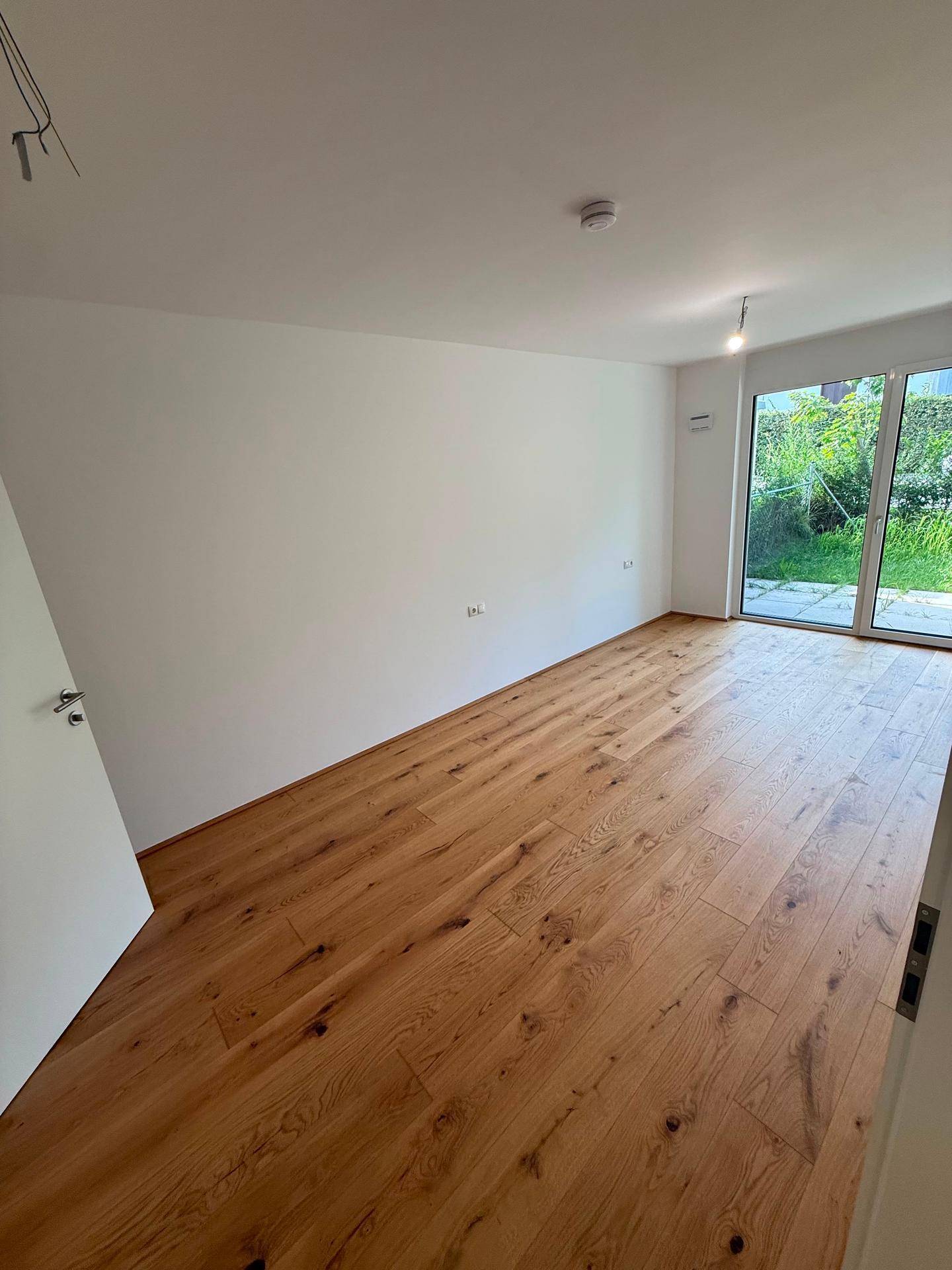 Leeres Zimmer mit Holzboden und großen Fenstern, die viel Tageslicht hereinlassen und einen Blick ins Grüne bieten.