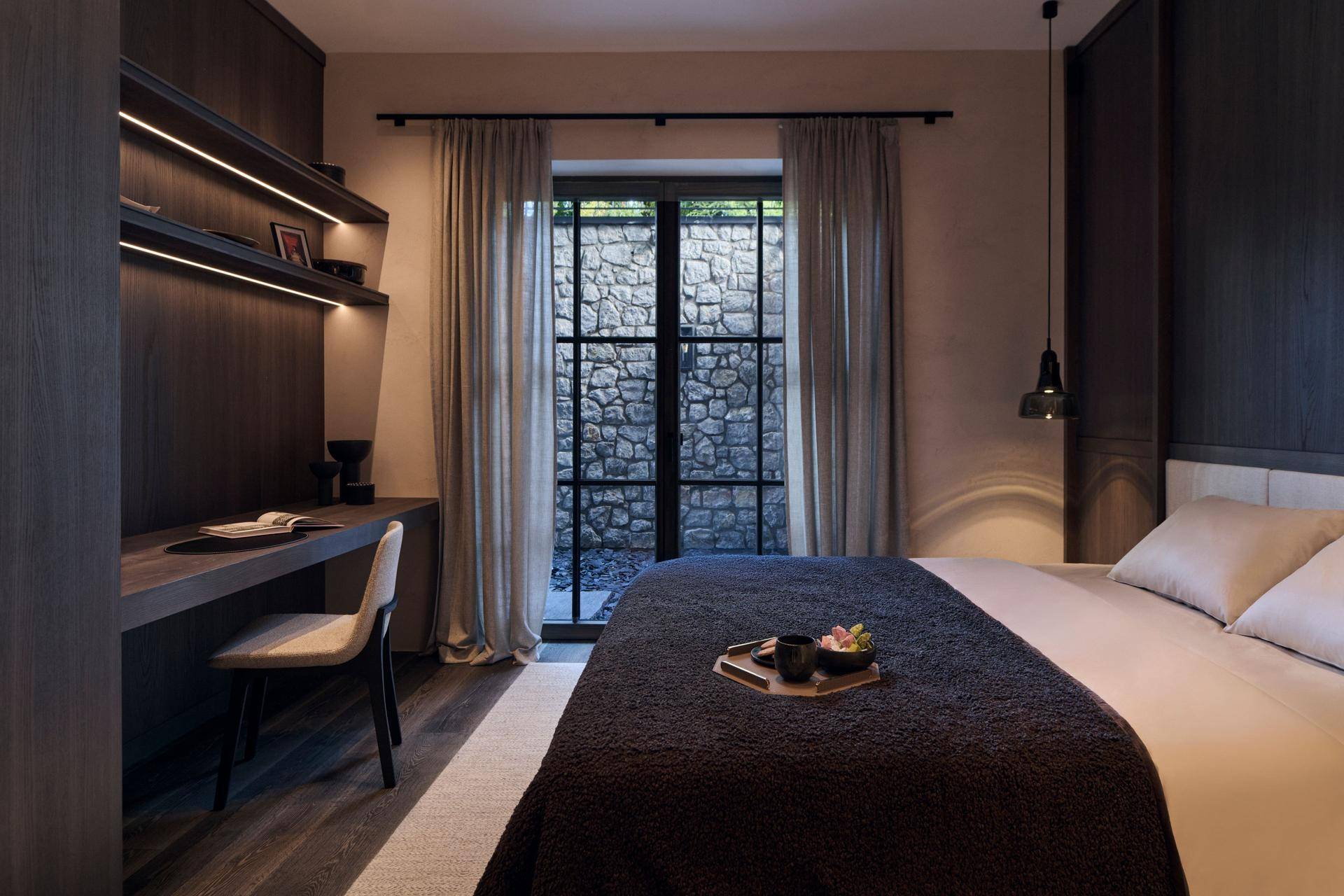 Ruhiges Schlafzimmer mit Schreibtisch und Fensterblick auf eine Natursteinwand.